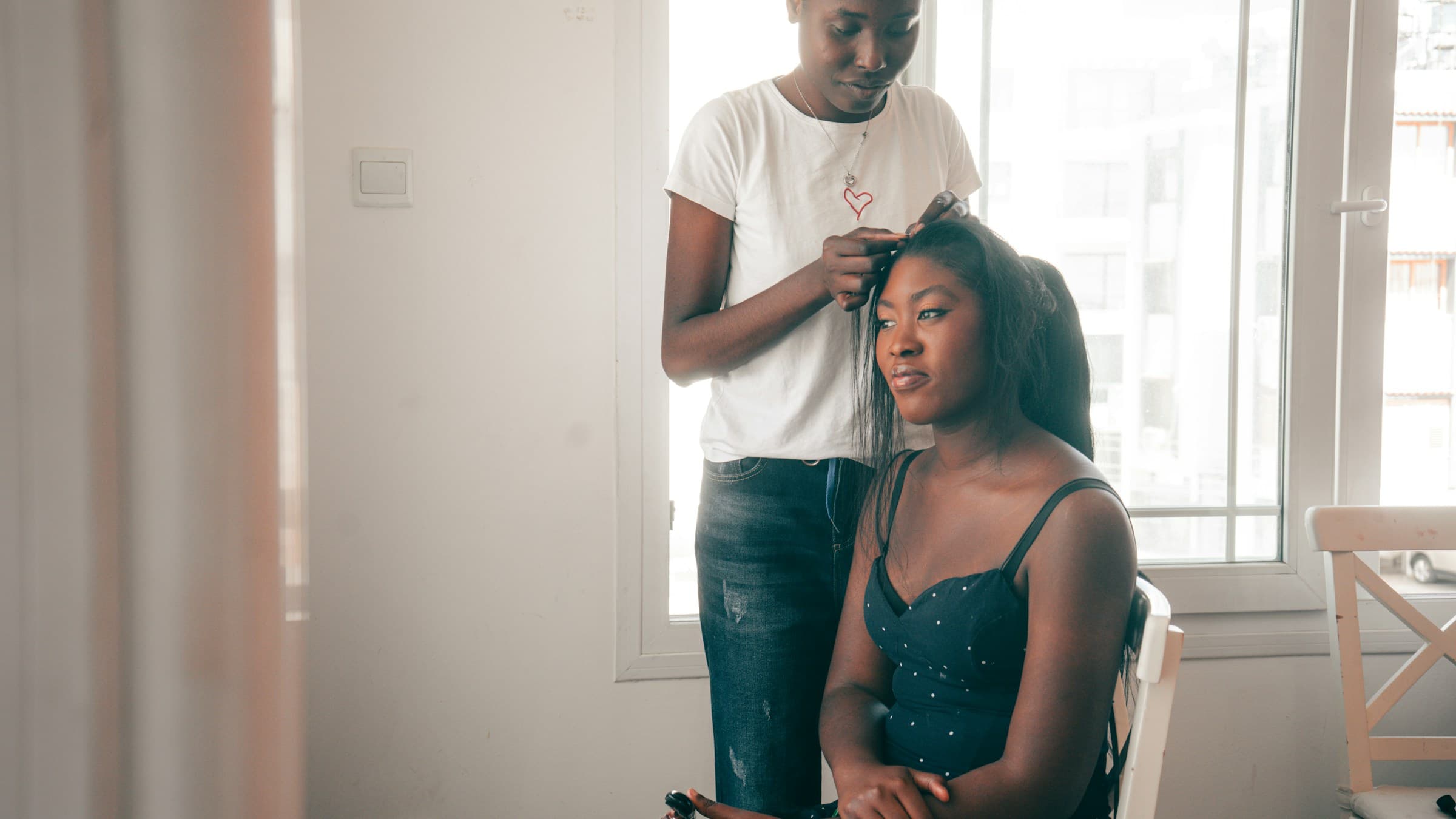 Cabeleireira faz um penteado em uma cliente sentada perto da janela