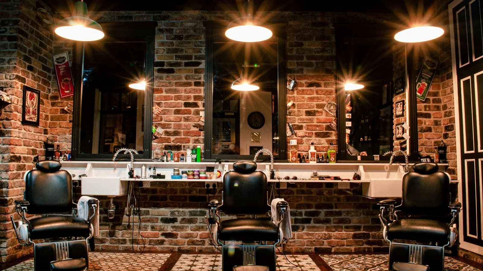 Três cadeiras de barbeiro vazias diante dos espelhos, numa barbearia de parede de tijolos