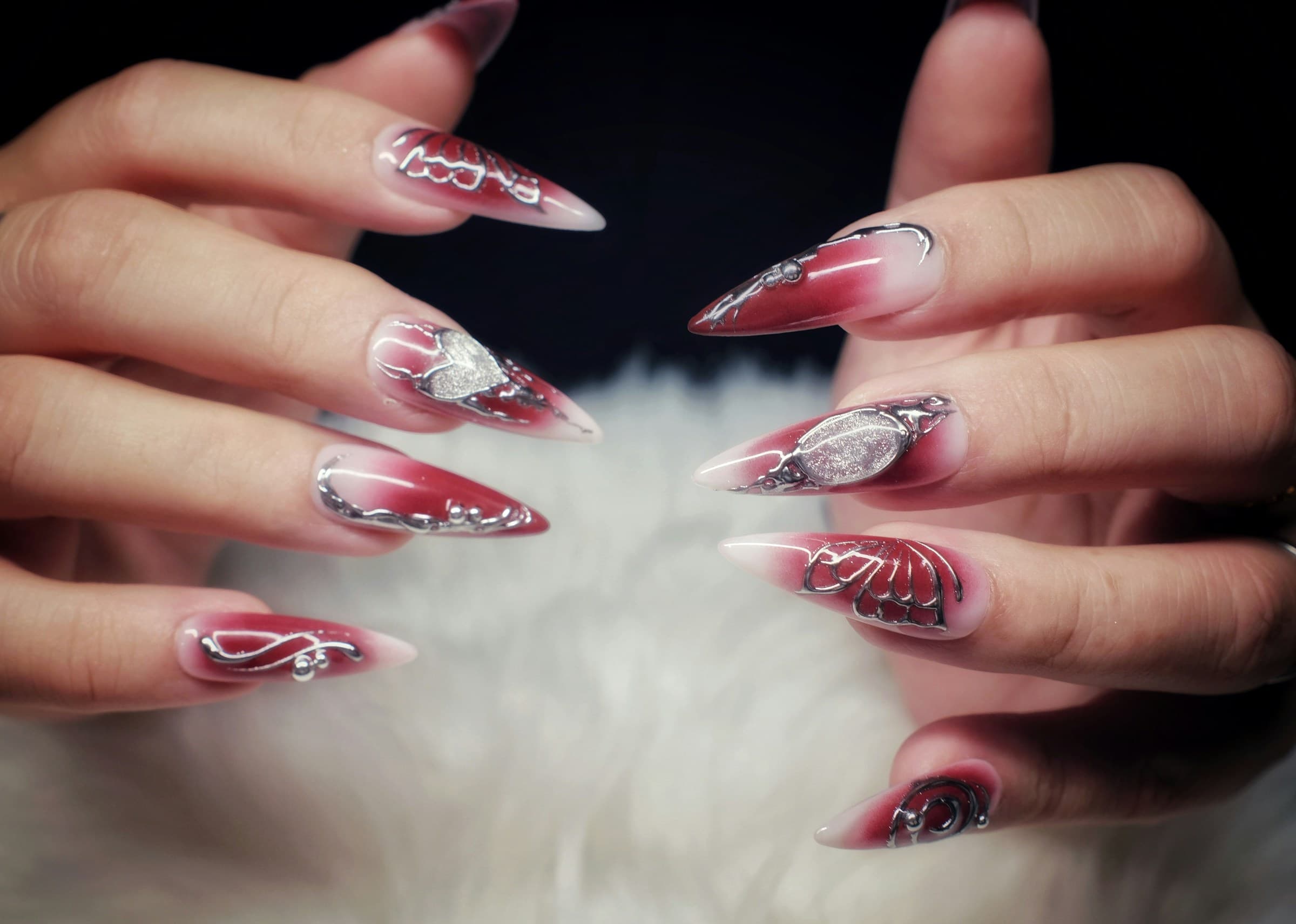 Mãos exibem unhas longas em stiletto com degradê vermelho e desenhos prateados sobre fundo de pelúcia