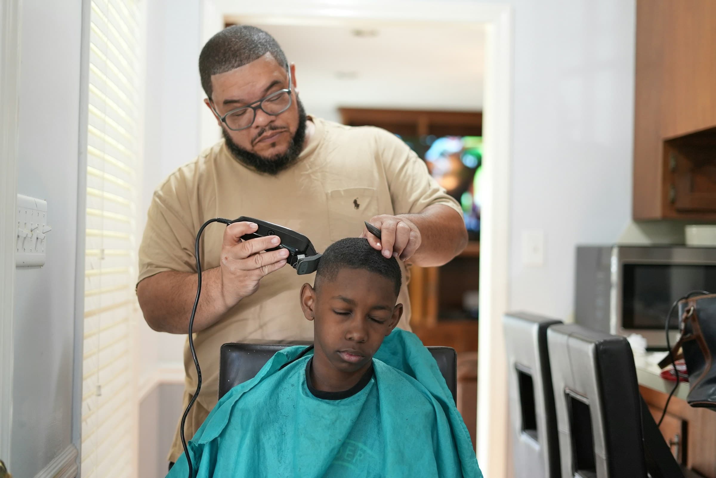 Barbeiro corta o cabelo de um menino com máquina dentro de casa, perto da cozinha