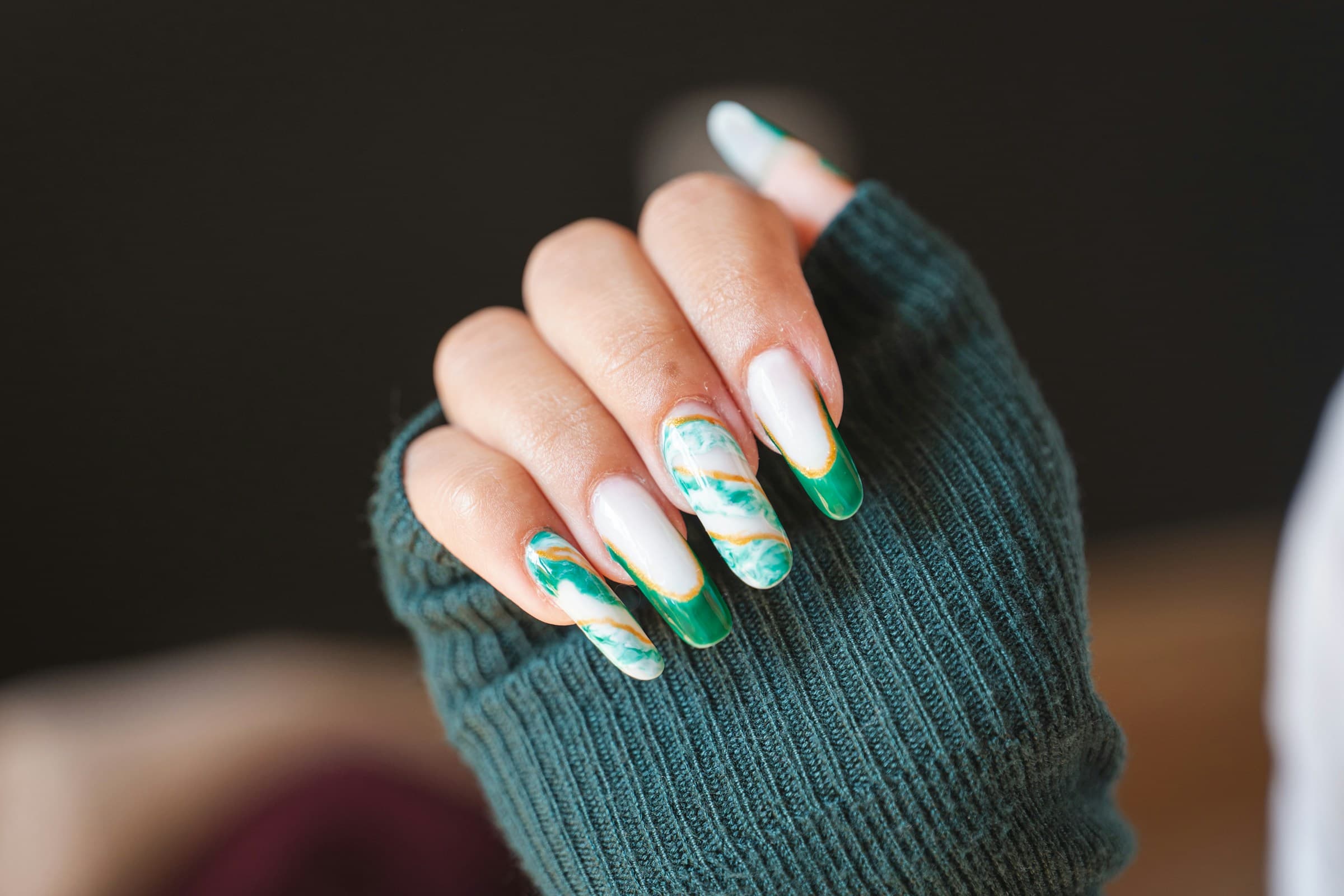 Mão com unhas longas amendoadas decoradas em mármore verde e branco, sobre suéter verde