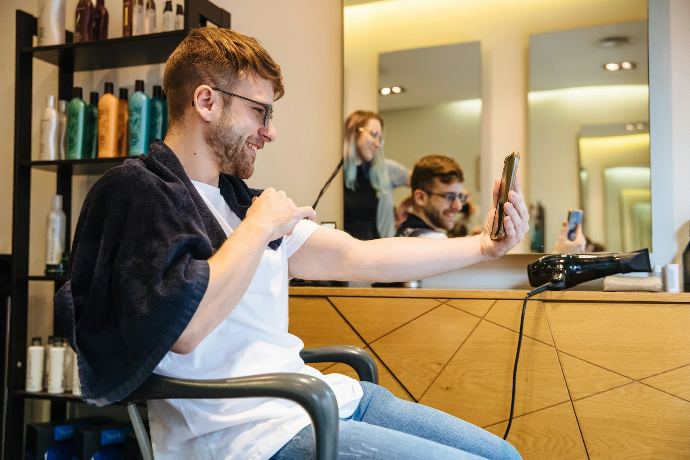 Cliente de barba tira uma selfie com o celular depois do corte, sentado na cadeira em frente ao espelho