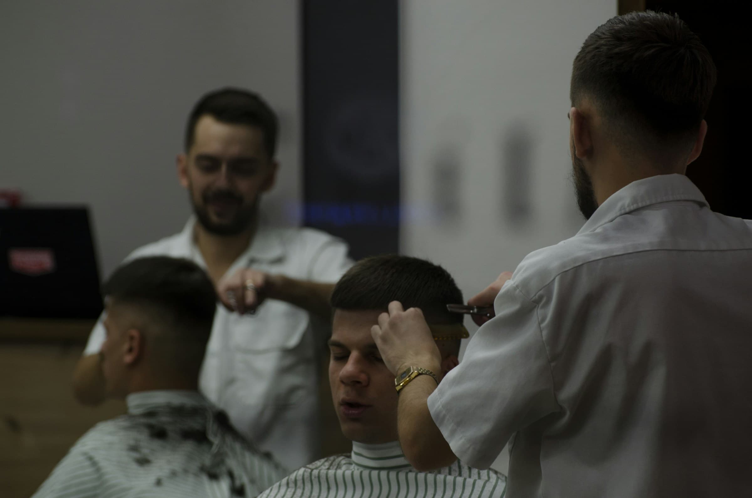 Barbeiro jovem corta o cabelo de um cliente enquanto outro barbeiro trabalha ao lado