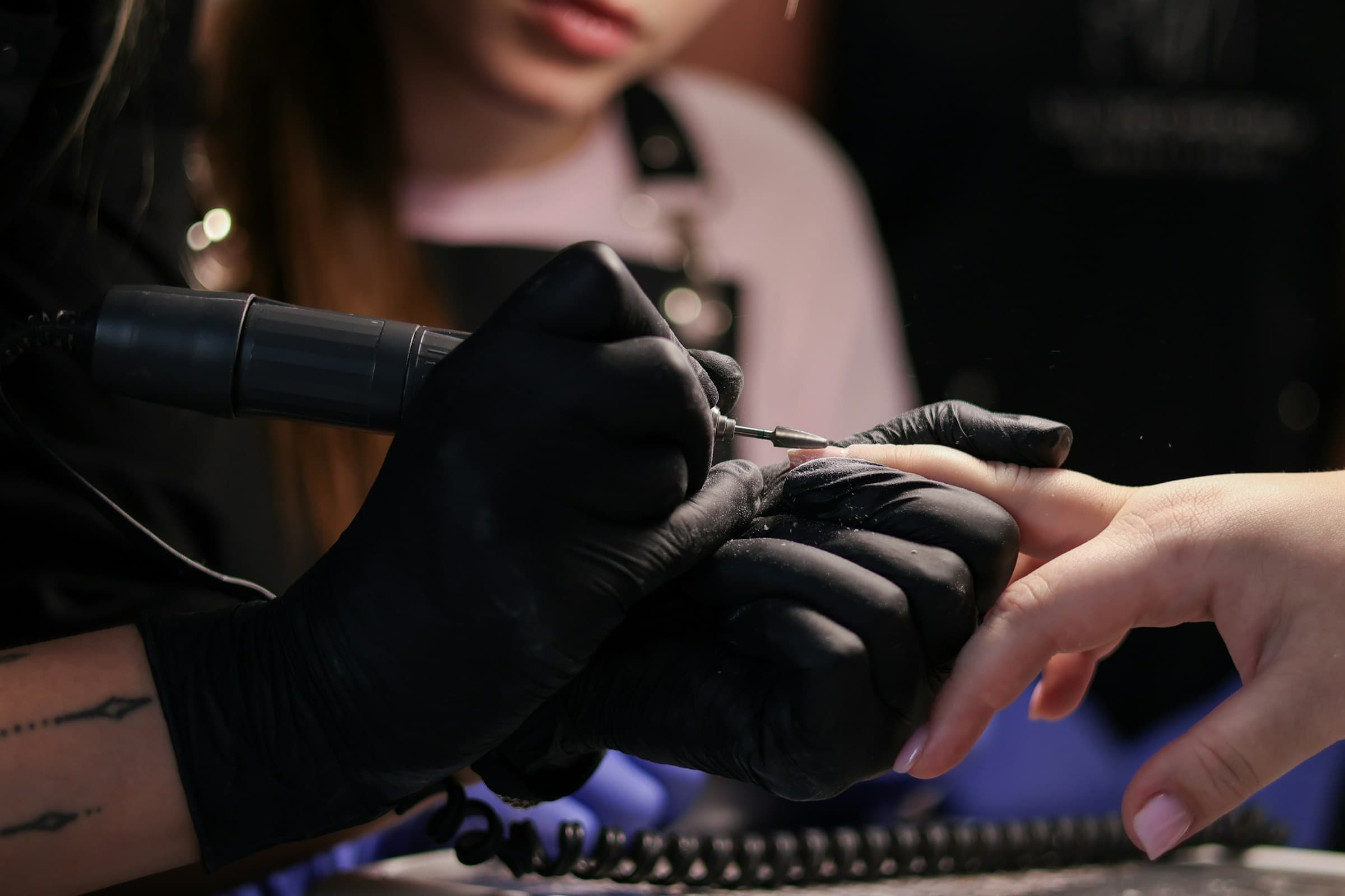 Manicure de luvas pretas usa a lixa elétrica na manutenção das unhas de uma cliente
