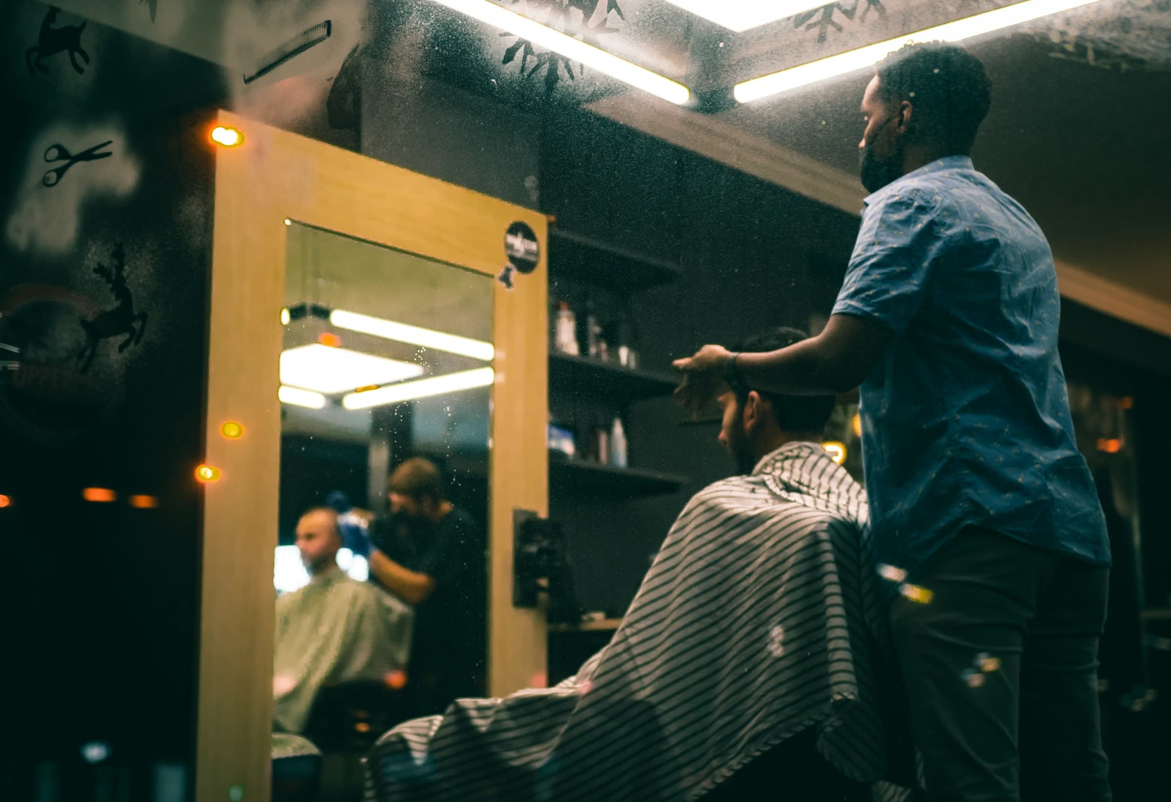 Barbeiro de camisa azul atende um cliente na cadeira diante de um espelho iluminado
