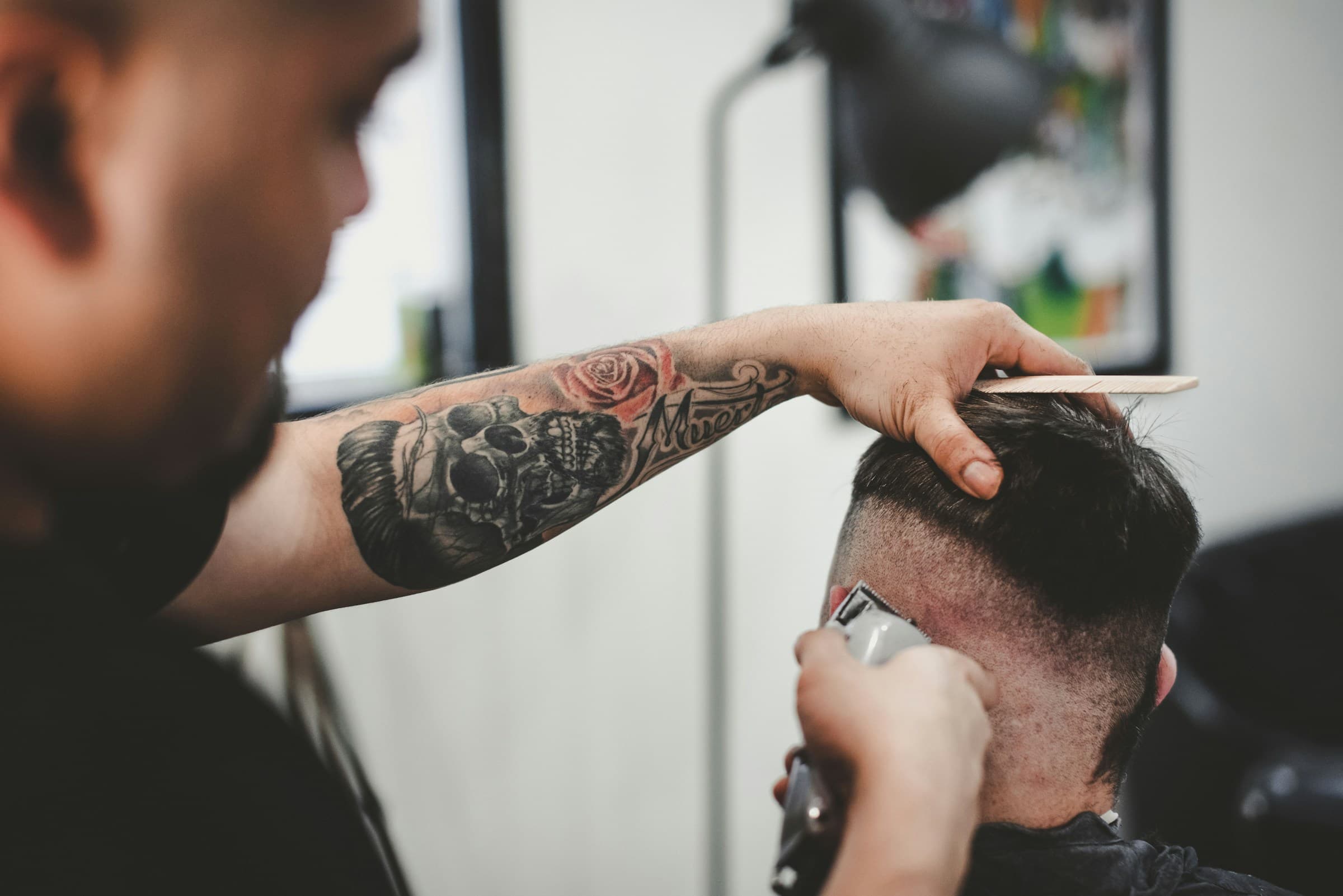Barbeiro tatuado faz um degradê com máquina e pente na nuca do cliente