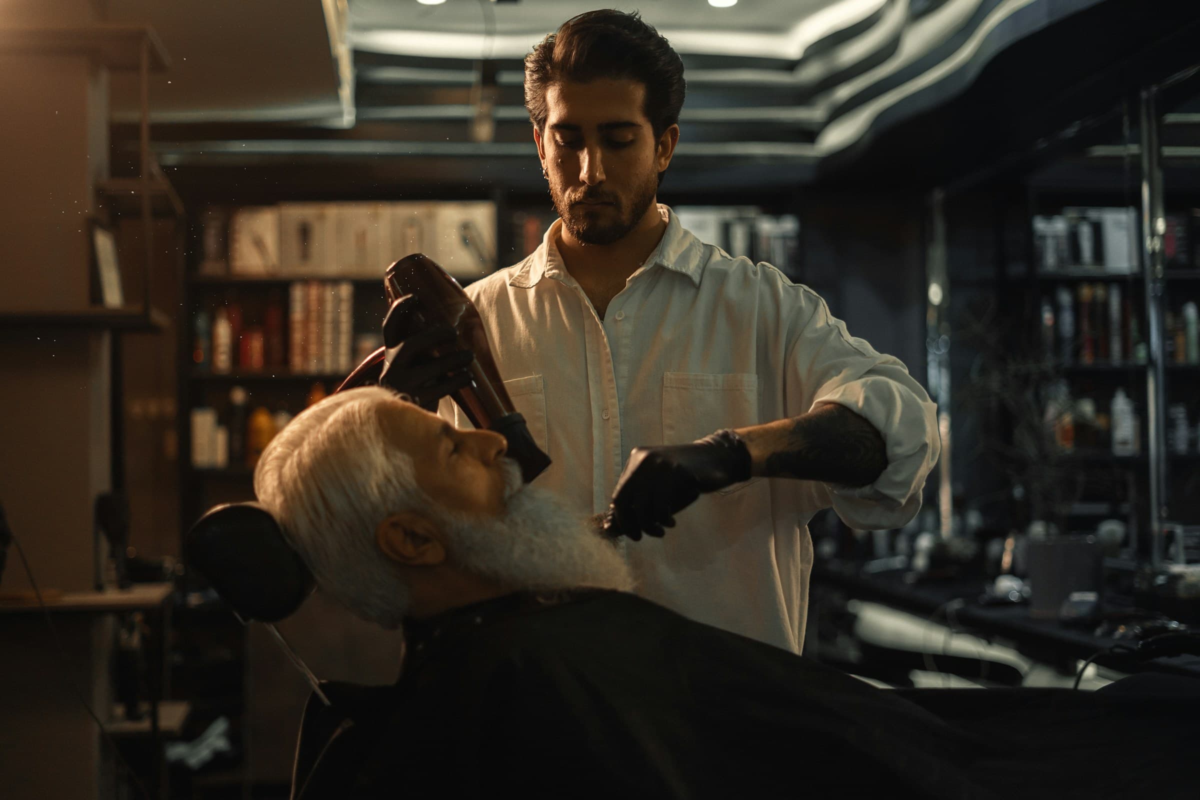 Barbeiro finaliza o cabelo de um cliente de barba branca na cadeira, em barbearia de luz baixa